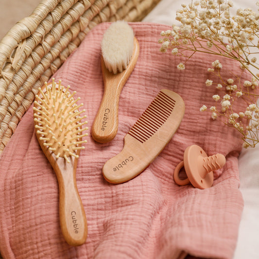 a set of three hairbrushes are laid on a pink muslin blanket with a pacifier on the side